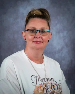 Portrait of a woman with light brown hair in a top bun, wearing turquoise glasses and a white graphic t-shirt, facing the camera against a blue-gray backdrop.