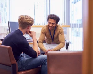 Two men sit on chairs in a bright office, smiling and talking during a one-on-one session.